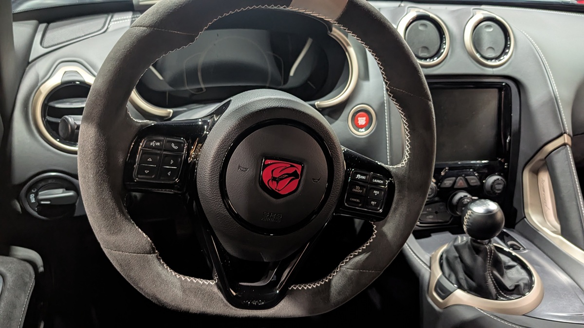 Close-up view of a car’s interior, showing a black leather-wrapped steering wheel with a red snake logo, a manual gear shift, dashboard controls, and circular air vents.