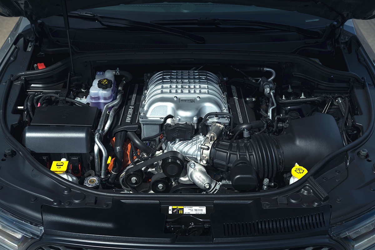 A close-up view of a car engine bay, showing a large supercharged V8 engine with various components, belts, and fluid reservoirs clearly visible under the hood.