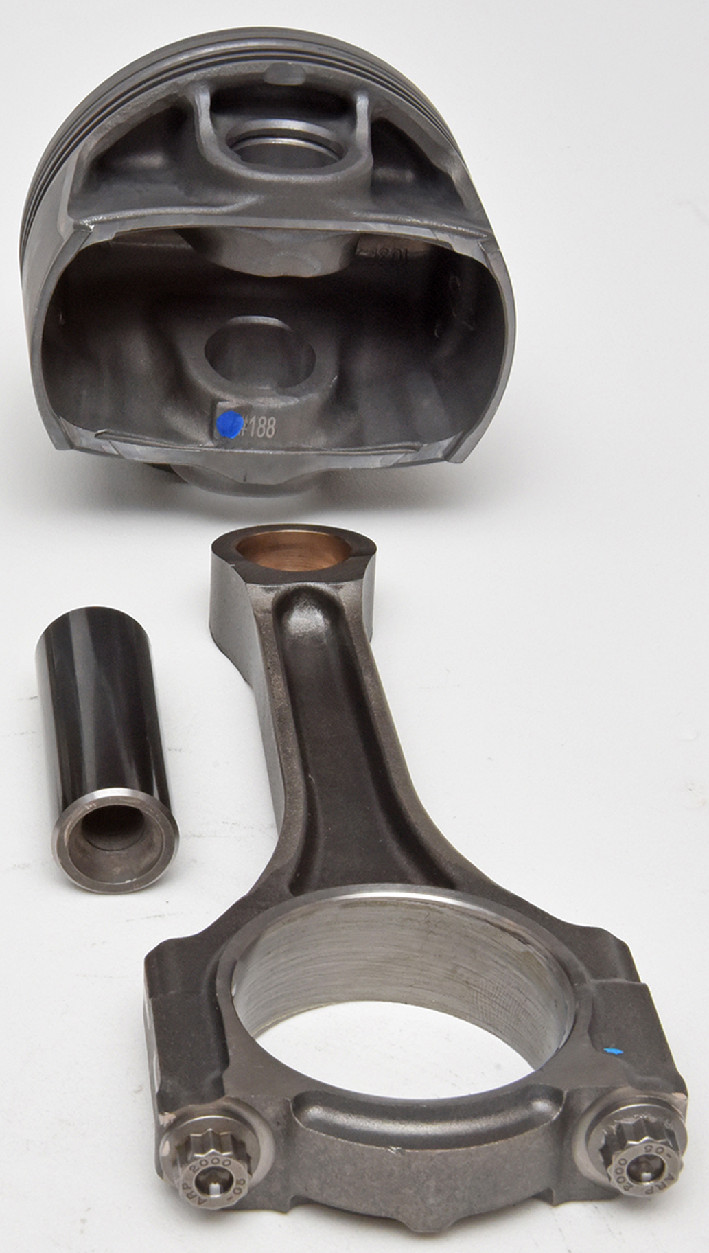 A metal piston, connecting rod, and piston pin from an engine are arranged on a white background, showing the different components separated from each other.