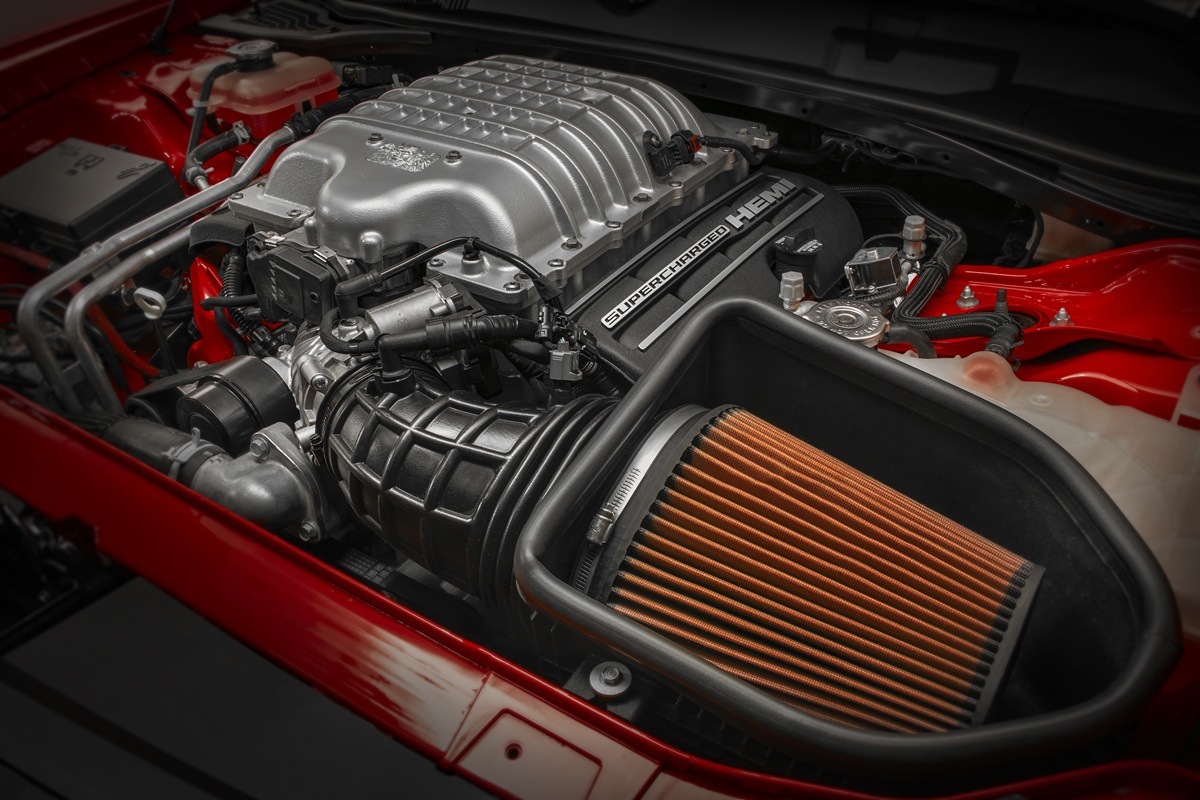 Close-up view of a supercharged HEMI engine inside a car, featuring a prominent air intake with an orange filter, metallic components, hoses, and parts of the red car body visible around the engine bay.
