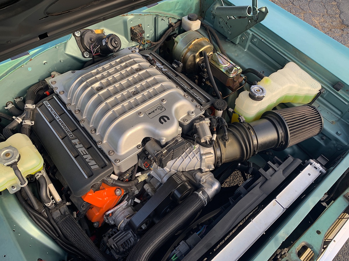 Close-up view of a supercharged HEMI V8 engine installed in a vintage car with a teal engine bay, showing detailed components like air intake, coolant reservoir, and hoses.