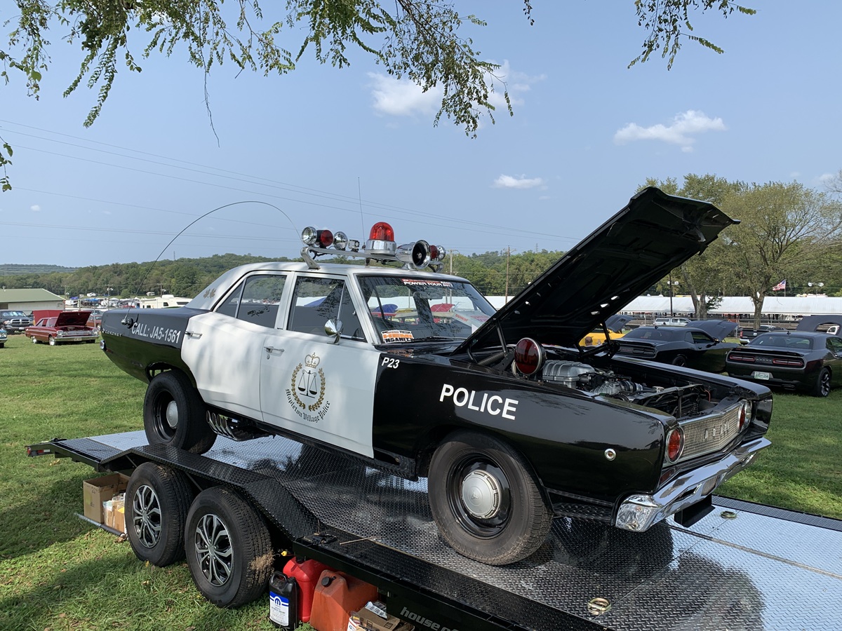 A vintage black and white police car with its hood open is displayed on a trailer outdoors, surrounded by grass, trees, and parked cars under a clear sky.