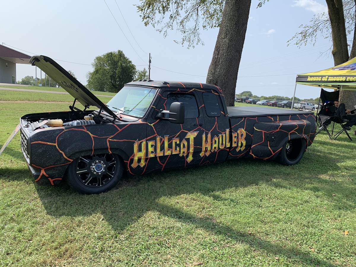A customized black pickup truck with an orange cracked-lava paint design and the words HELLCAT HAULER on the side is parked on grass with its hood open at an outdoor event.