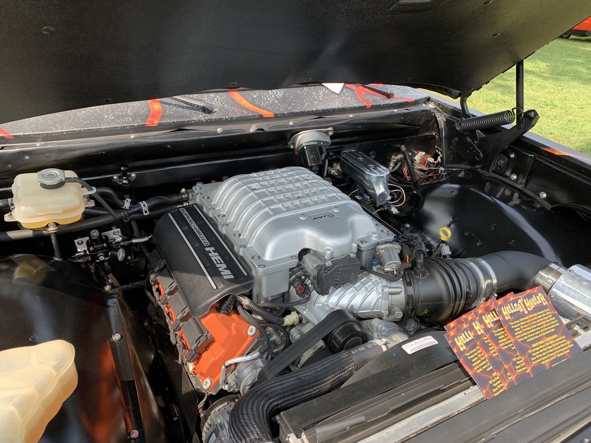 A close-up view of a supercharged HEMI engine inside a car’s open hood, showing various engine components, wires, and fluid reservoirs, with grass visible outside the vehicle.