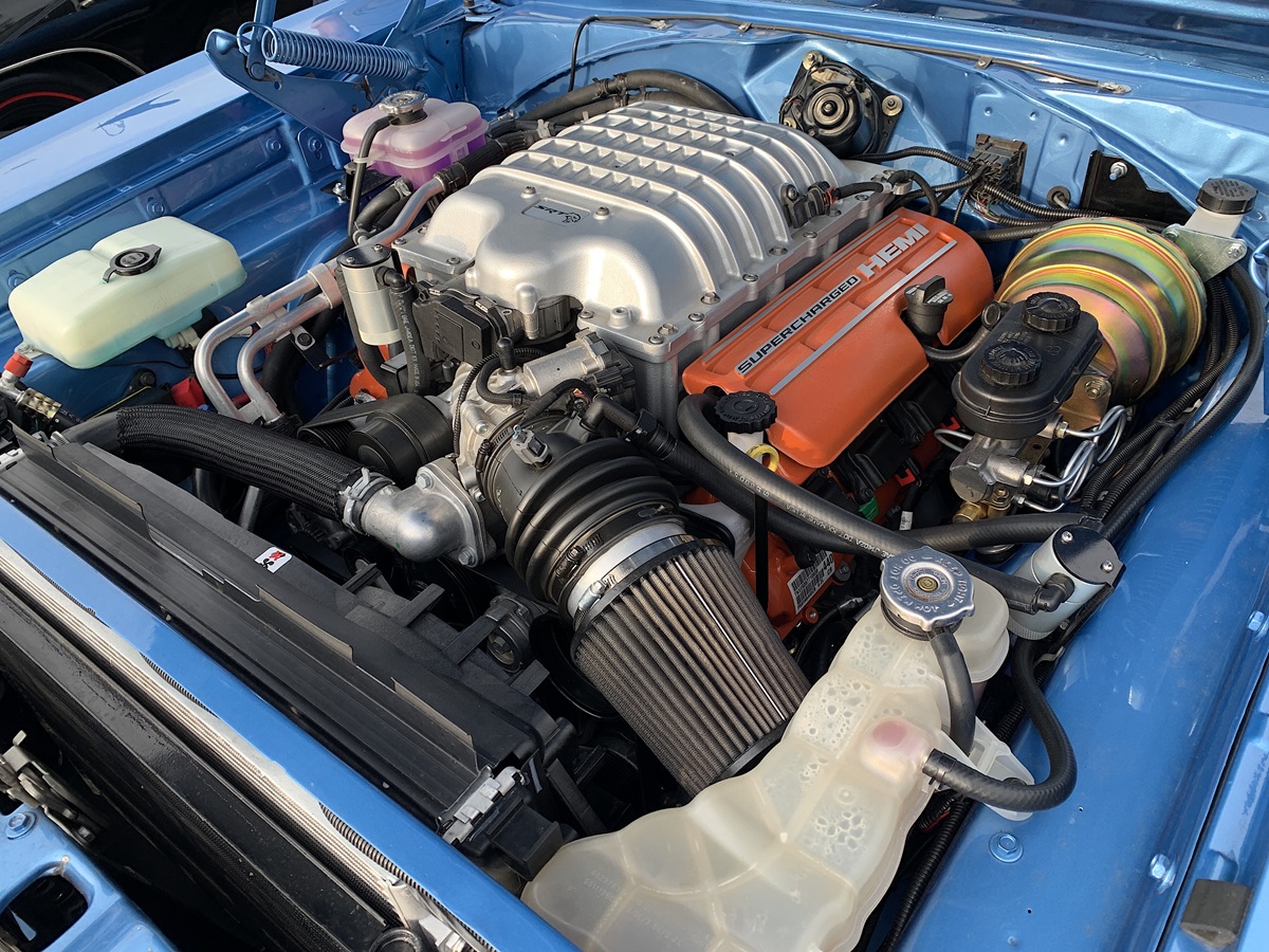 A detailed view of a car engine bay showing a modern supercharged HEMI V8 engine with orange valve covers installed in a classic blue car. The components and hoses are clean and well-organized.