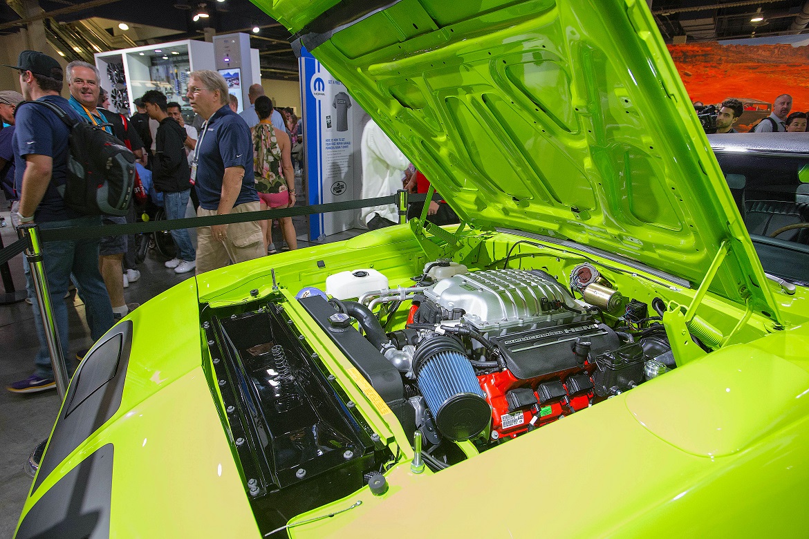 A bright green classic car with its hood open displays a modified engine at an indoor auto show. Several people stand around, admiring the vehicle in a busy exhibition hall.