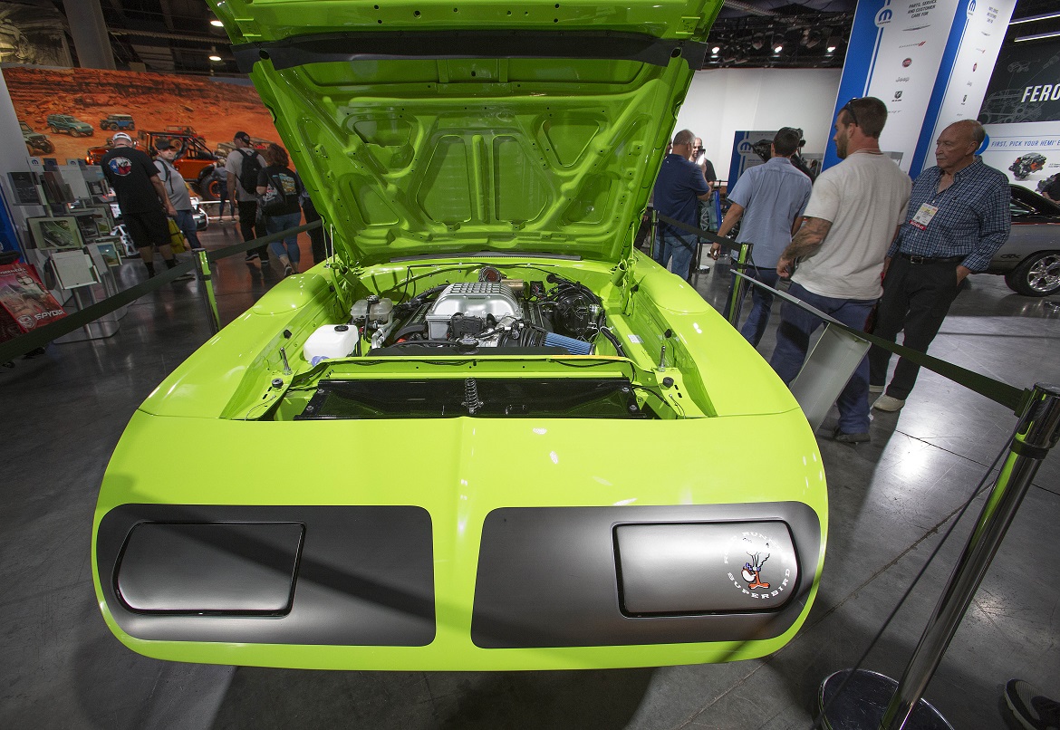 Neon green classic muscle car with its hood open, showcasing the engine at an indoor auto show. Several people are observing the car, and exhibits and banners are visible in the background.