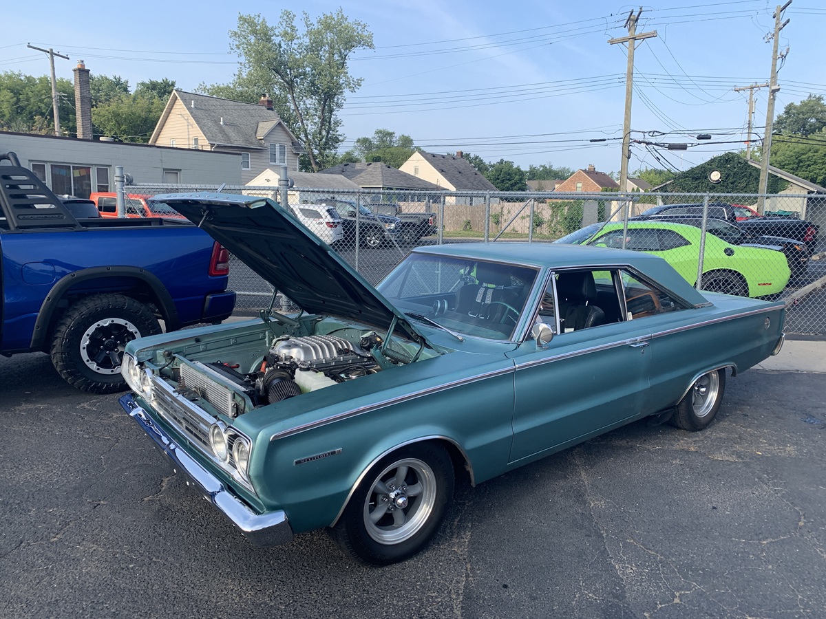 A classic teal muscle car with its hood open is parked on pavement near a chain-link fence. Other vehicles, houses, and power lines are visible in the background.