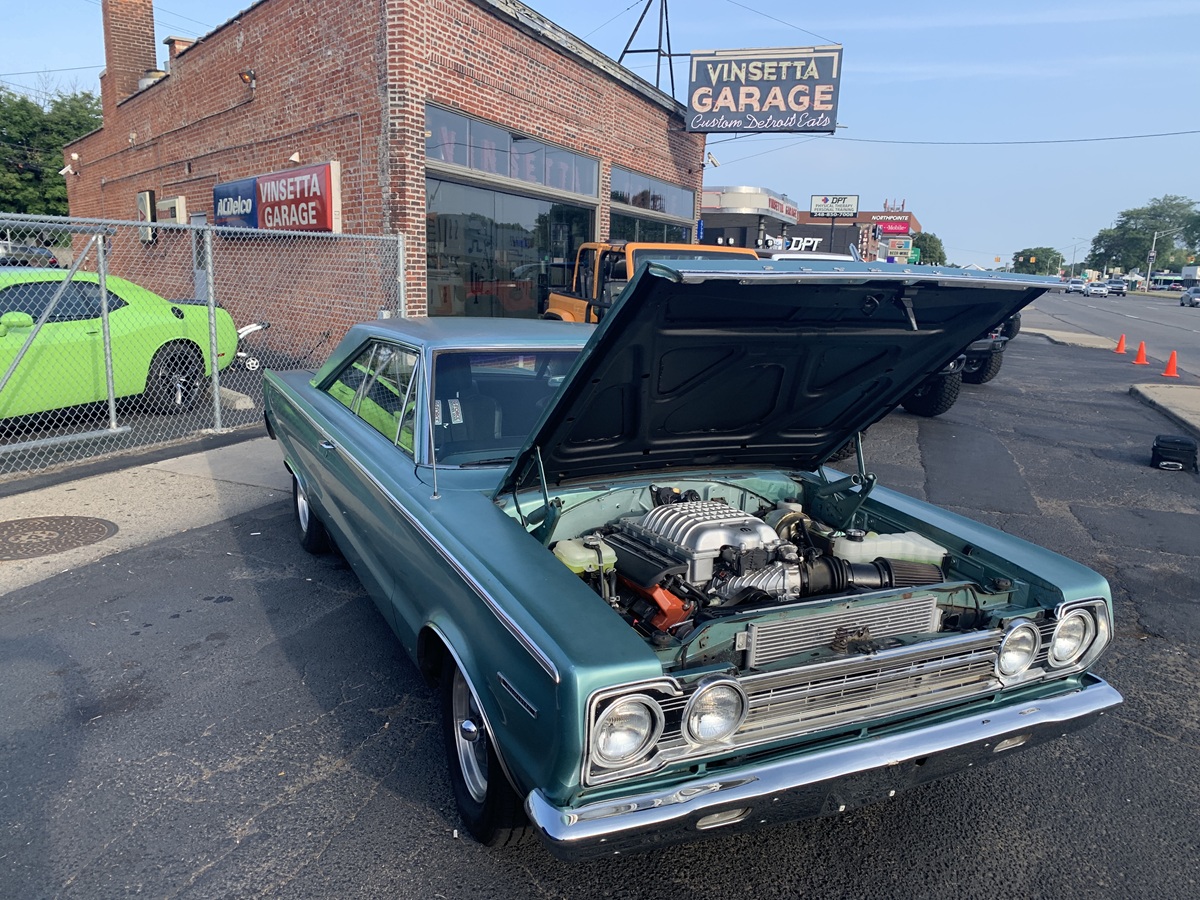 A classic teal Plymouth car with its hood open, revealing the engine, is parked outside Vinsetta Garage, a brick auto shop, on a sunny day. Other cars and traffic cones are visible in the background.