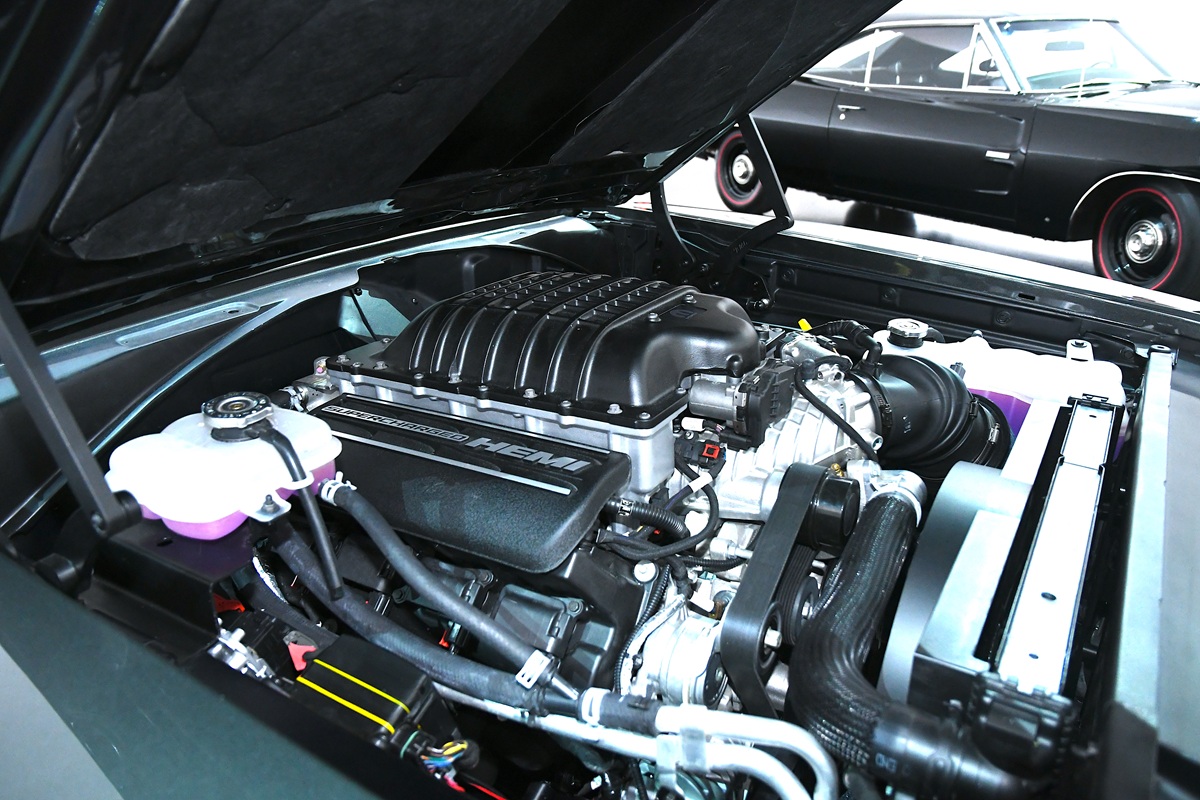 A close-up view of a supercharged HEMI engine under the hood of a car, with various engine components visible. Another classic car with red-rimmed tires is parked in the background.