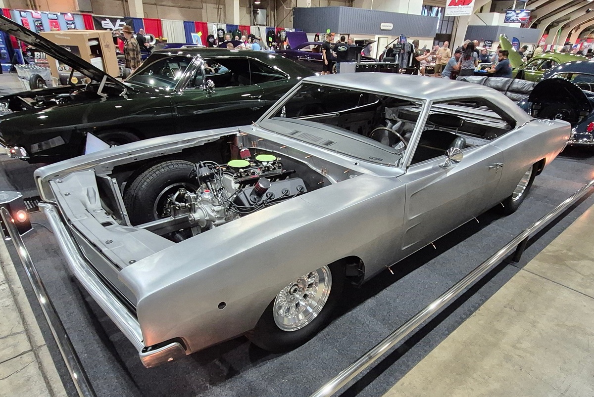 A silver classic muscle car with its hood removed, revealing the engine, is displayed indoors at a car show among other vintage vehicles. The car appears unfinished, with no interior or front grille.