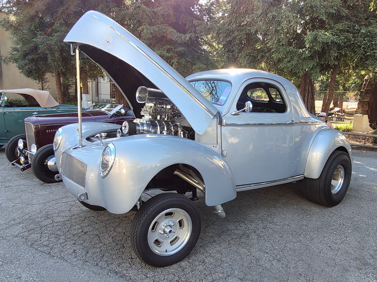 A light gray classic hot rod car with its front hood open, showcasing its engine. The car is parked outdoors on a paved area, with trees and other vintage cars in the background.