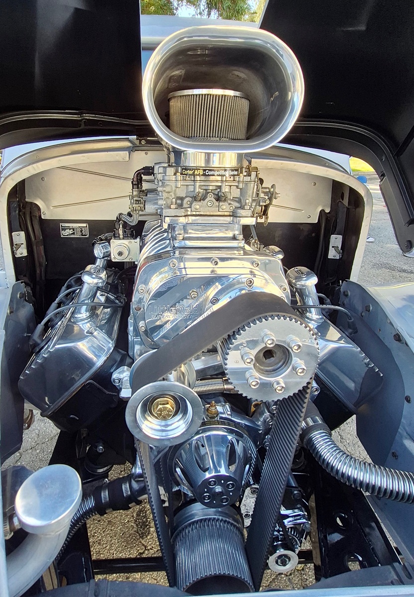 Close-up view of a highly polished, chrome-plated supercharged car engine with a large air intake scoop and exposed pulleys and belt, situated inside a classic car’s engine bay.