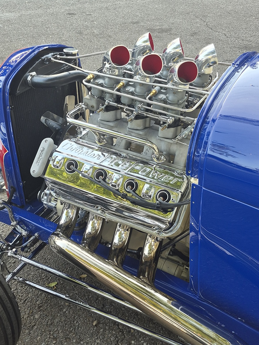 A close-up of a shiny chrome V8 engine with exposed headers and four carburetors in a blue hot rod, parked on an asphalt surface.