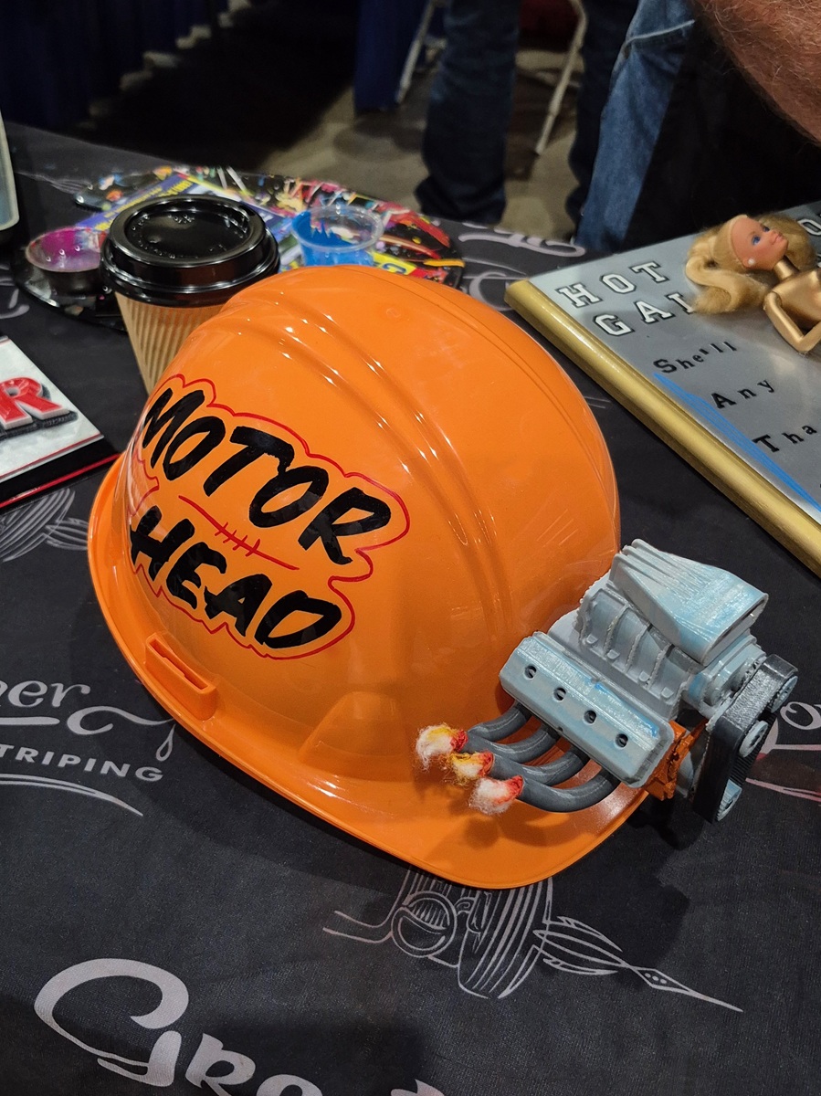An orange hardhat with MOTOR HEAD written on it and a small model engine attached to the side sits on a table alongside a coffee cup, book, and other assorted items.
