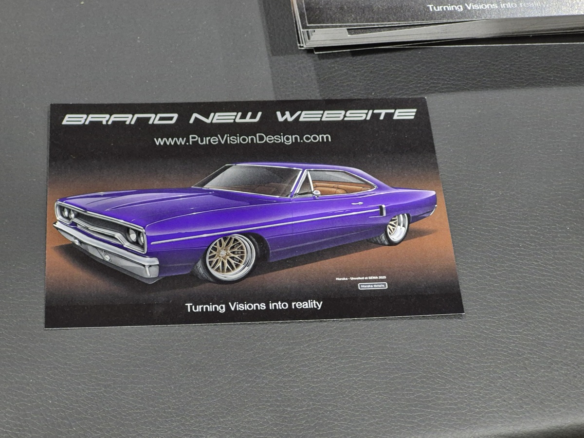 A flyer featuring a purple classic muscle car illustration, promoting a new website for PureVisionDesign.com with the tagline Turning Visions into reality. The flyer is placed on a gray surface.