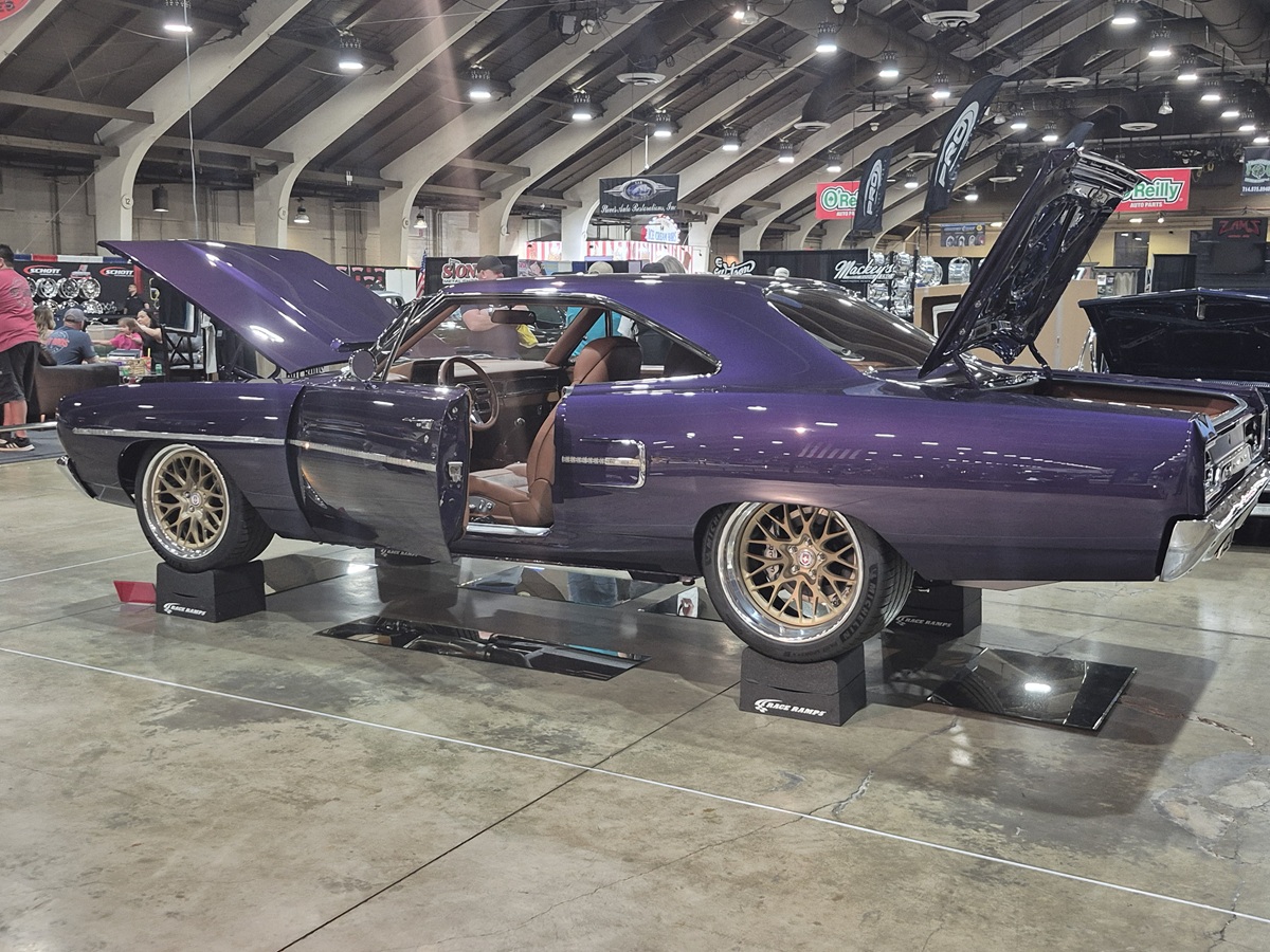 A restored purple classic muscle car is displayed indoors with its hood, doors, and trunk open, showcasing its interior and engine at an auto show. The car is raised on platforms with bright overhead lighting.