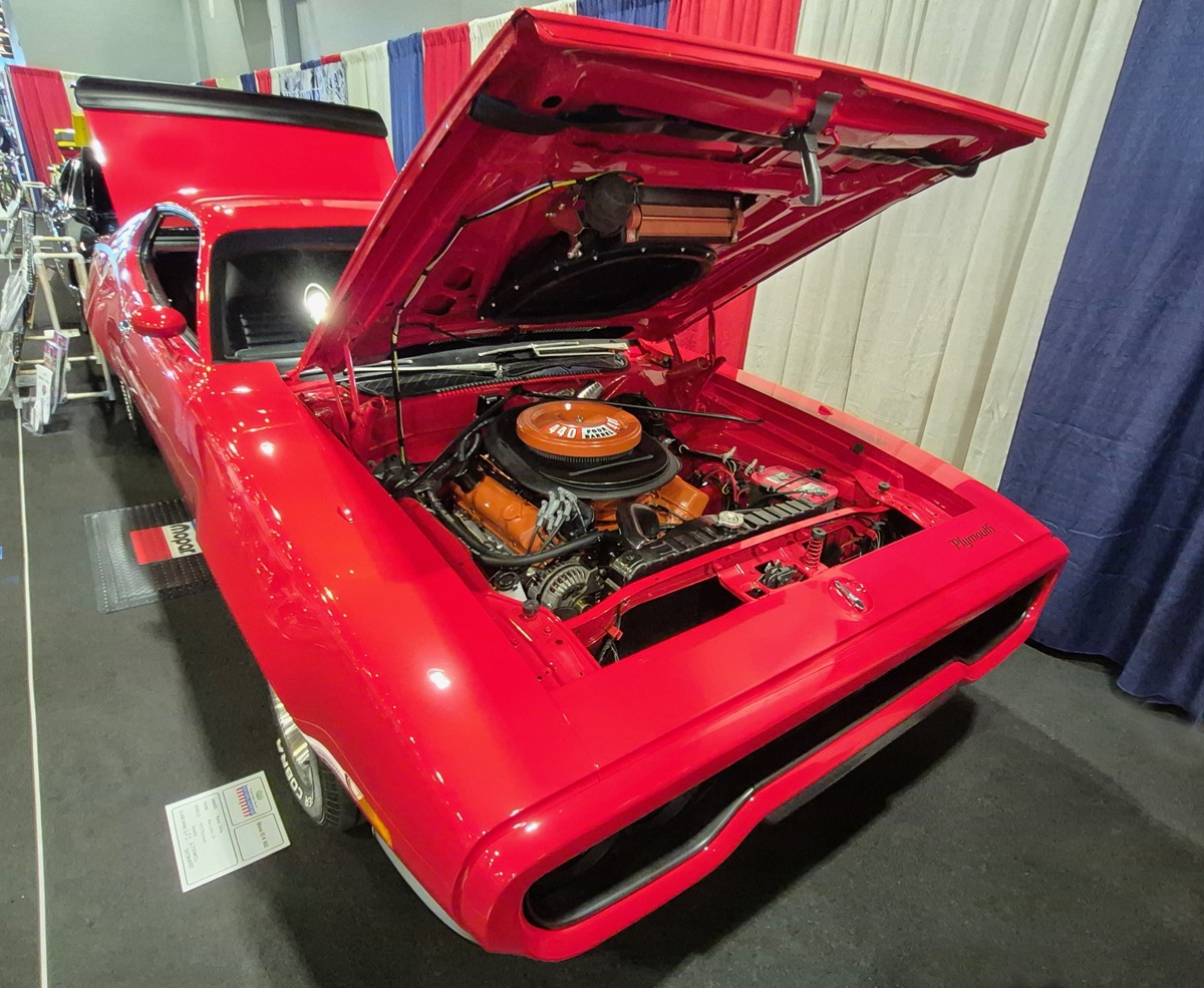 A bright red classic muscle car with its hood open, showcasing a clean, powerful engine. The car is displayed indoors with red, white, and blue curtains in the background.