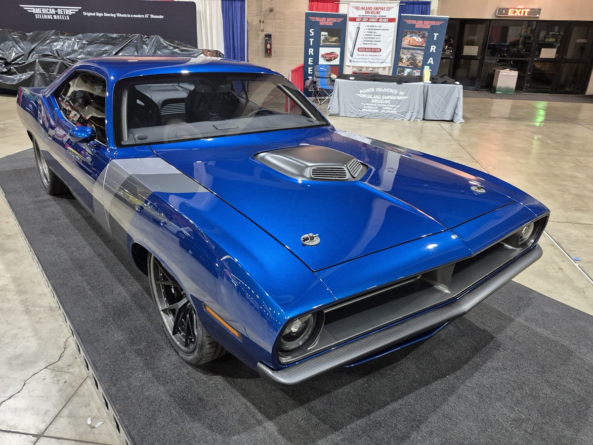 A bright blue classic muscle car with a hood scoop on display indoors, parked on a gray mat with promotional banners and tables in the background.