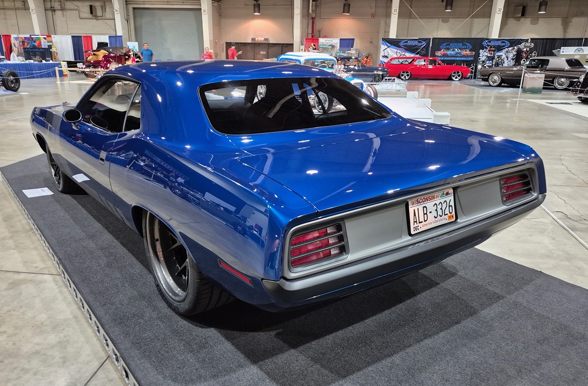 A blue classic muscle car with black wheels is displayed indoors at a car show. The car is on a dark carpet, with other vintage cars and display booths visible in the background.