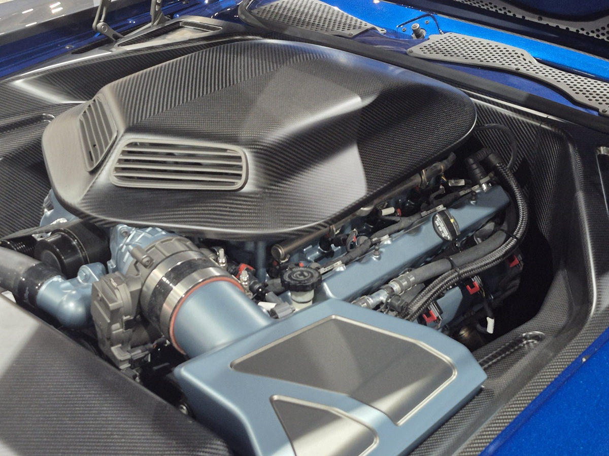 Close-up of a high-performance car engine with carbon fiber components and blue accents, showcasing intricate wiring and parts inside a blue sports car engine bay.