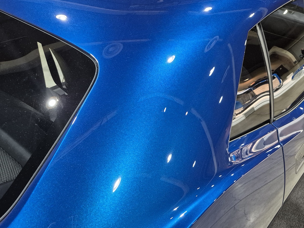 Close-up of the rear side of a shiny blue car, showing reflections of lights on the smooth, metallic paint surface and part of the window and interior.