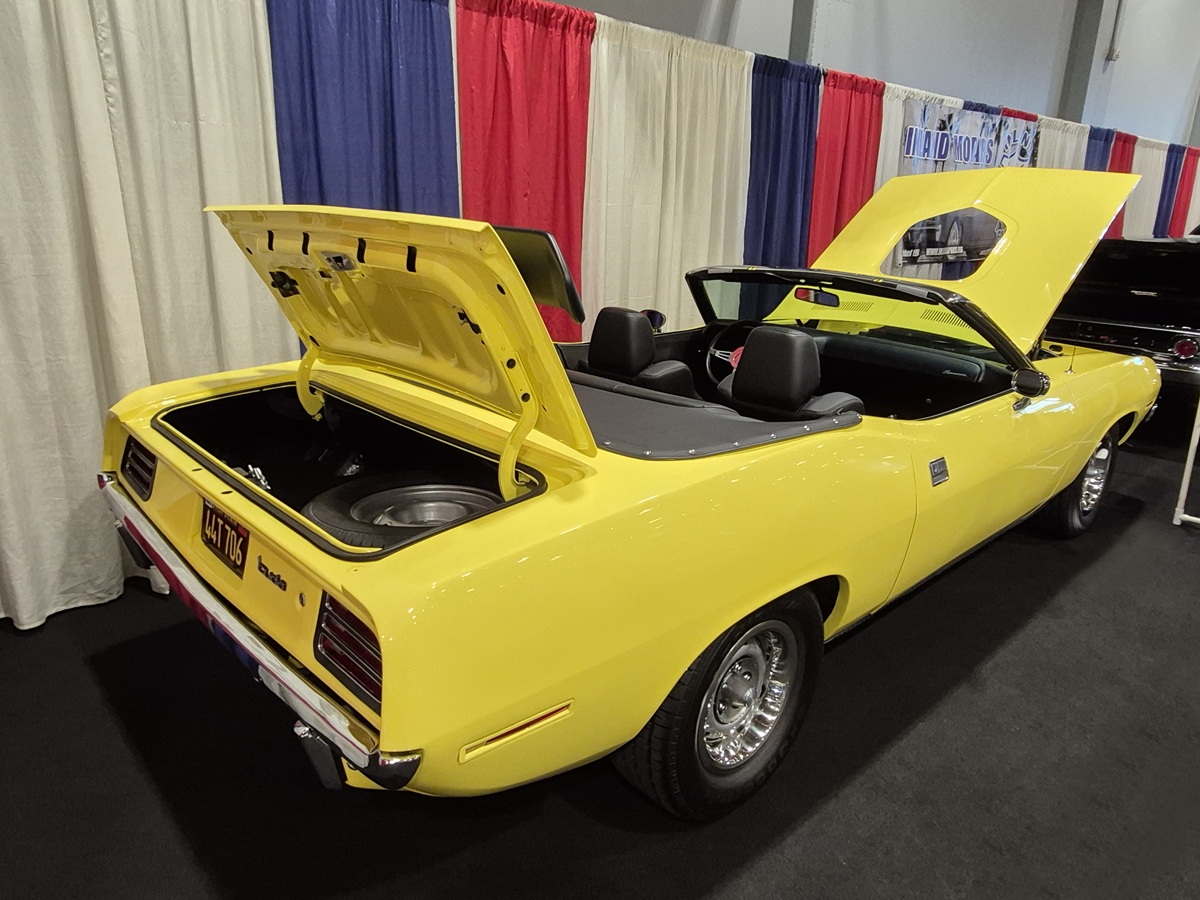 A bright yellow classic convertible car with its trunk and hood open is displayed indoors, against a backdrop of red, white, and blue curtains. The car features black interior and chrome wheels.