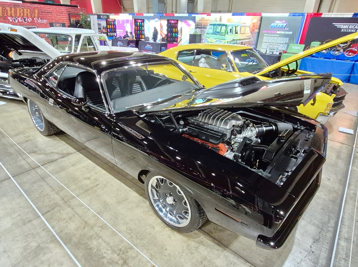 A shiny black classic car with its hood open, displaying its engine at an indoor car show. Another yellow classic car is parked beside it, with colorful displays in the background.