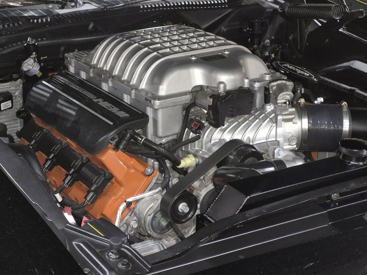 Close-up view of a supercharged car engine with a prominent silver supercharger on top, black intake tubing, orange valve covers, and various engine components visible.