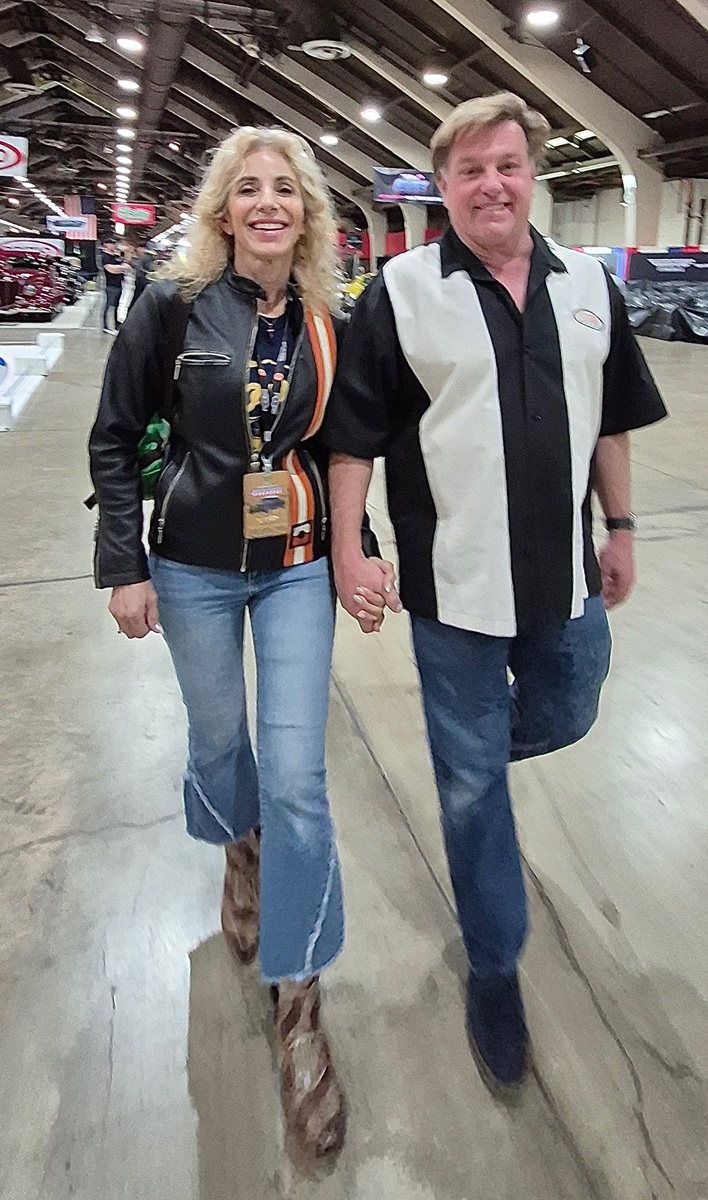 A smiling woman and man holding hands walk indoors at an event, wearing casual clothes—she in a black jacket and boots, he in a black and white shirt and jeans. The background shows an exhibition hall with booths and displays.