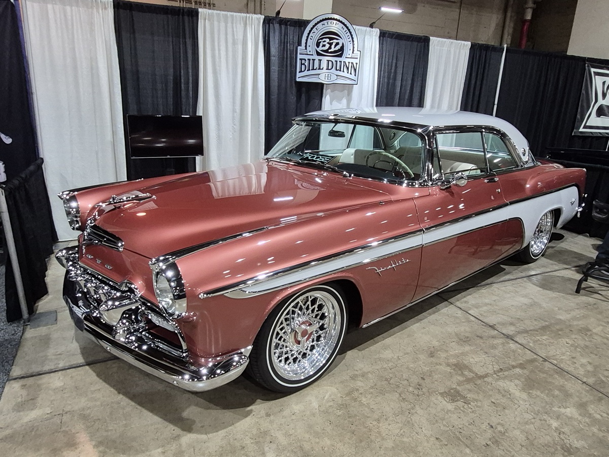 A classic two-tone pink and white Chrysler Imperial car is displayed indoors at a car show, with shiny chrome details and wire-spoke wheels, parked on a polished concrete floor.
