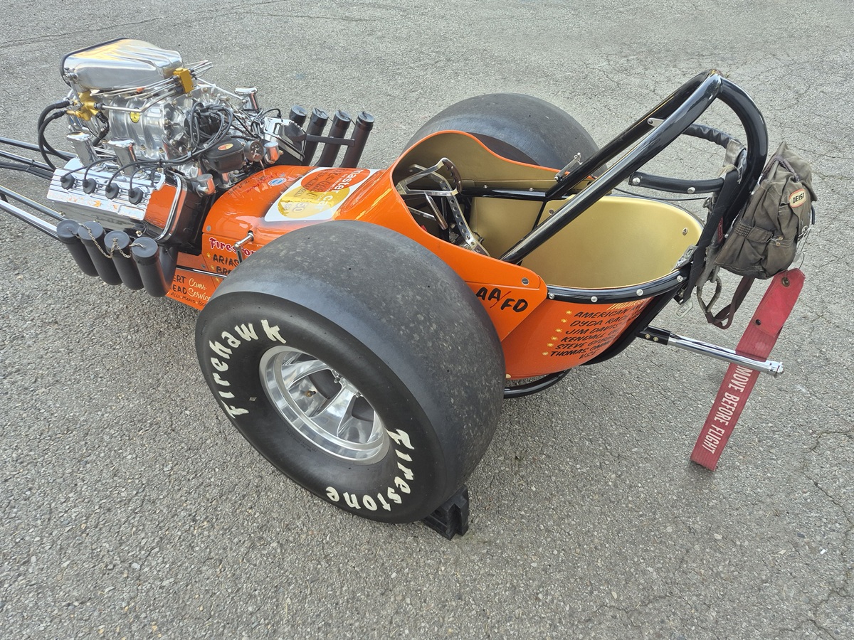 A close-up of a vintage orange dragster with large Firehawk rear tires, exposed engine, and a single seat. The car is parked on asphalt, and a brown bag hangs from the rear roll bar.