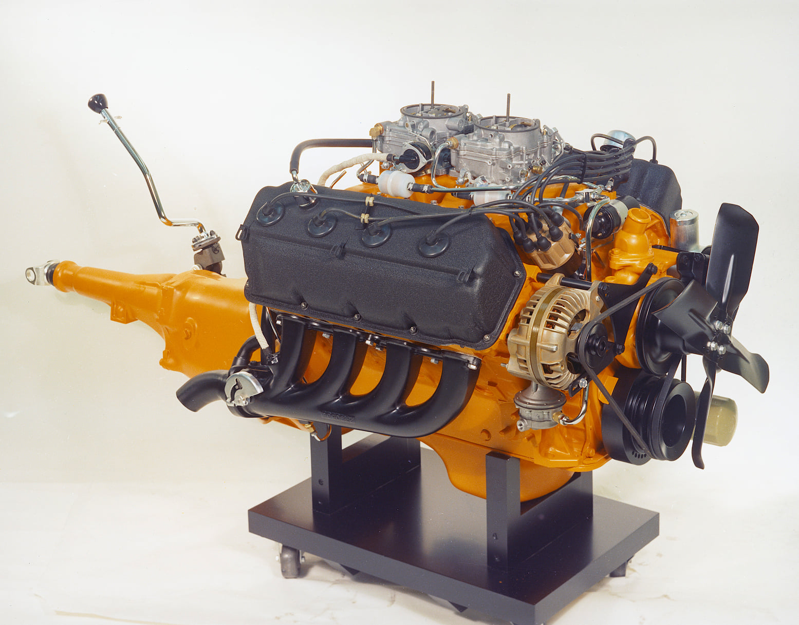 A vintage orange and black V8 car engine with visible transmission, carburetors, belts, fan, and exhaust headers, displayed on a black stand against a plain white background.