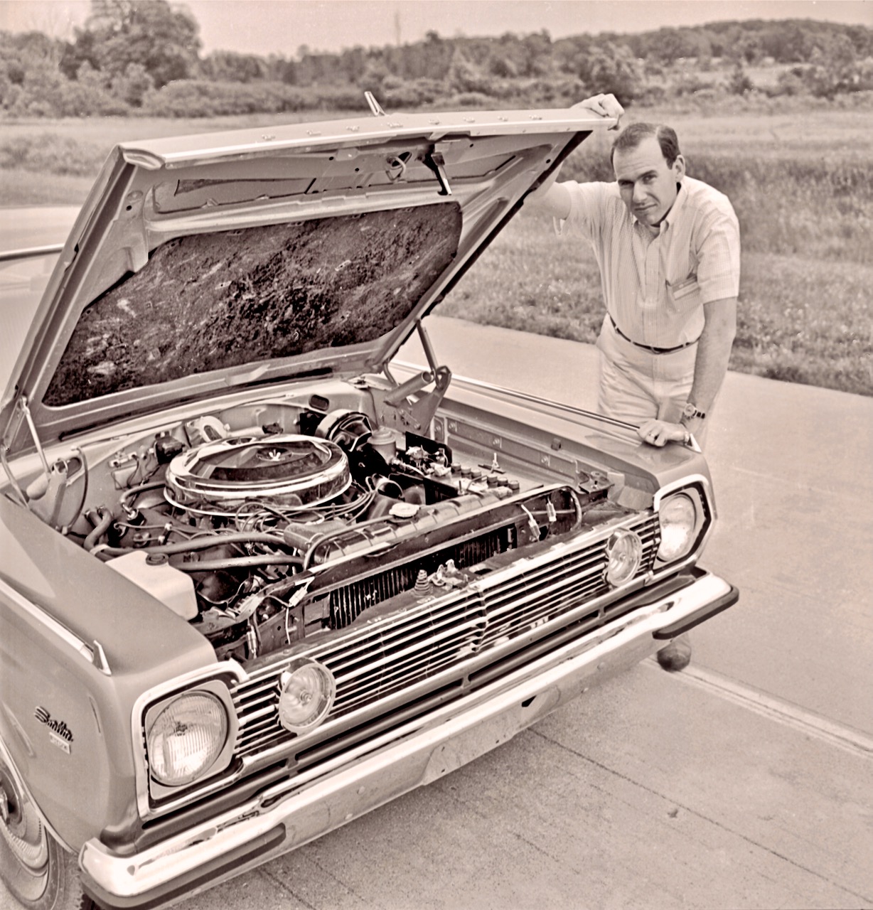 A man in light clothing leans on a classic car with an open hood, revealing its engine. The scene is outdoors on a country road with grassy fields and trees in the background. The image appears vintage or sepia-toned.
