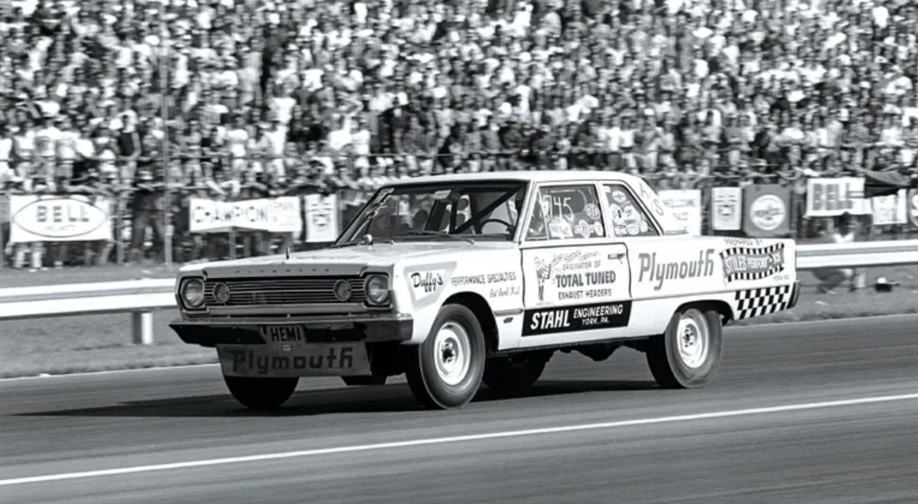A vintage Plymouth drag race car speeds down the track with sponsor decals on its side, in front of a large crowd of spectators in the stands. The photo is in black and white.