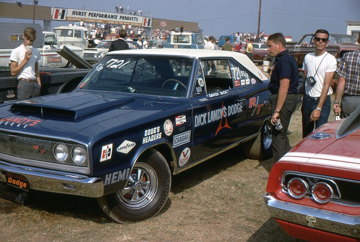 A vintage blue and white Dodge race car with Dick Landys Dodge on the side is parked at a car show; several men stand nearby and other classic cars are visible in the background.