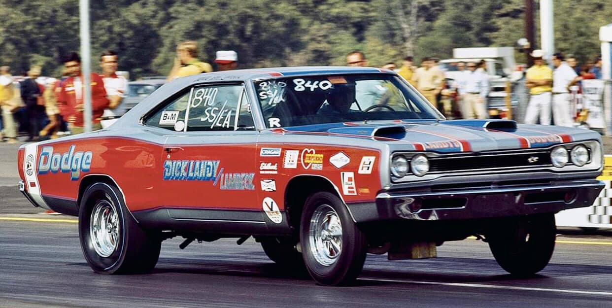 A red and blue classic Dodge drag racing car, with sponsor decals and raised front wheels, accelerates down a track as spectators and officials watch in the background.