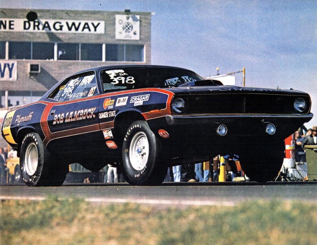 A classic drag racing car, a black and red Plymouth Barracuda, lifts its front wheels off the ground at the starting line of a dragstrip, with a building labeled DRAGWAY in the background.