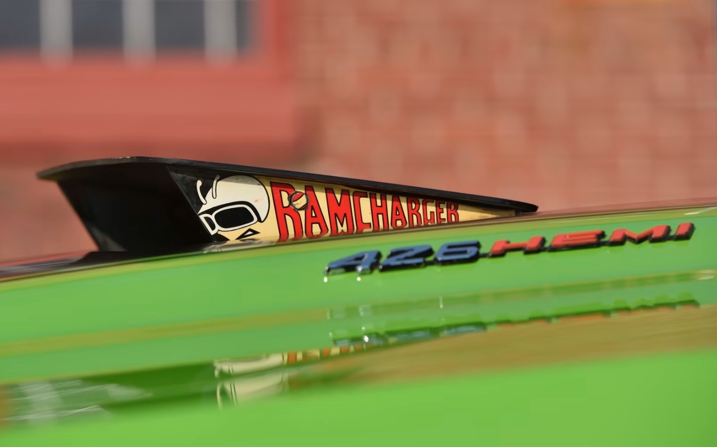 Close-up of a car hood featuring a 426 HEMI badge and a black hood scoop with a RAMCHARGER graphic showing a cartoon figure in a helmet, on a bright green vehicle.