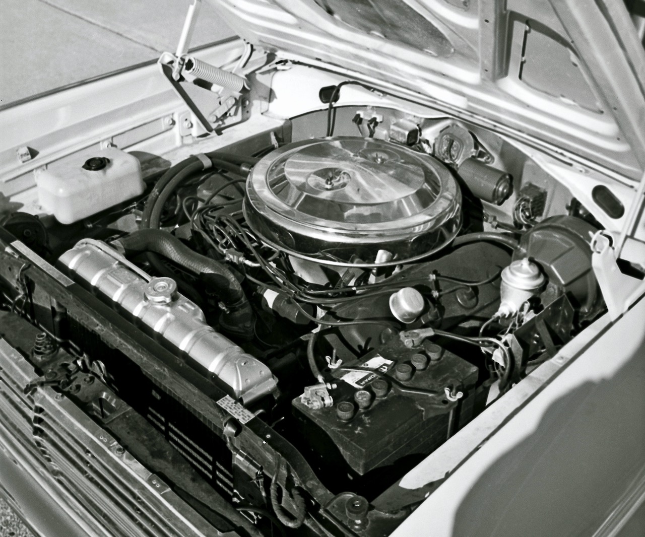 Black and white photo of a car engine under an open hood, showing various engine components, hoses, a large round air filter cover, radiator, and battery in a classic vehicle.