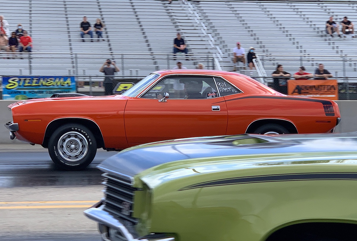 An orange classic muscle car races down a drag strip, with a green car partially visible in the foreground and empty bleachers with scattered spectators in the background.