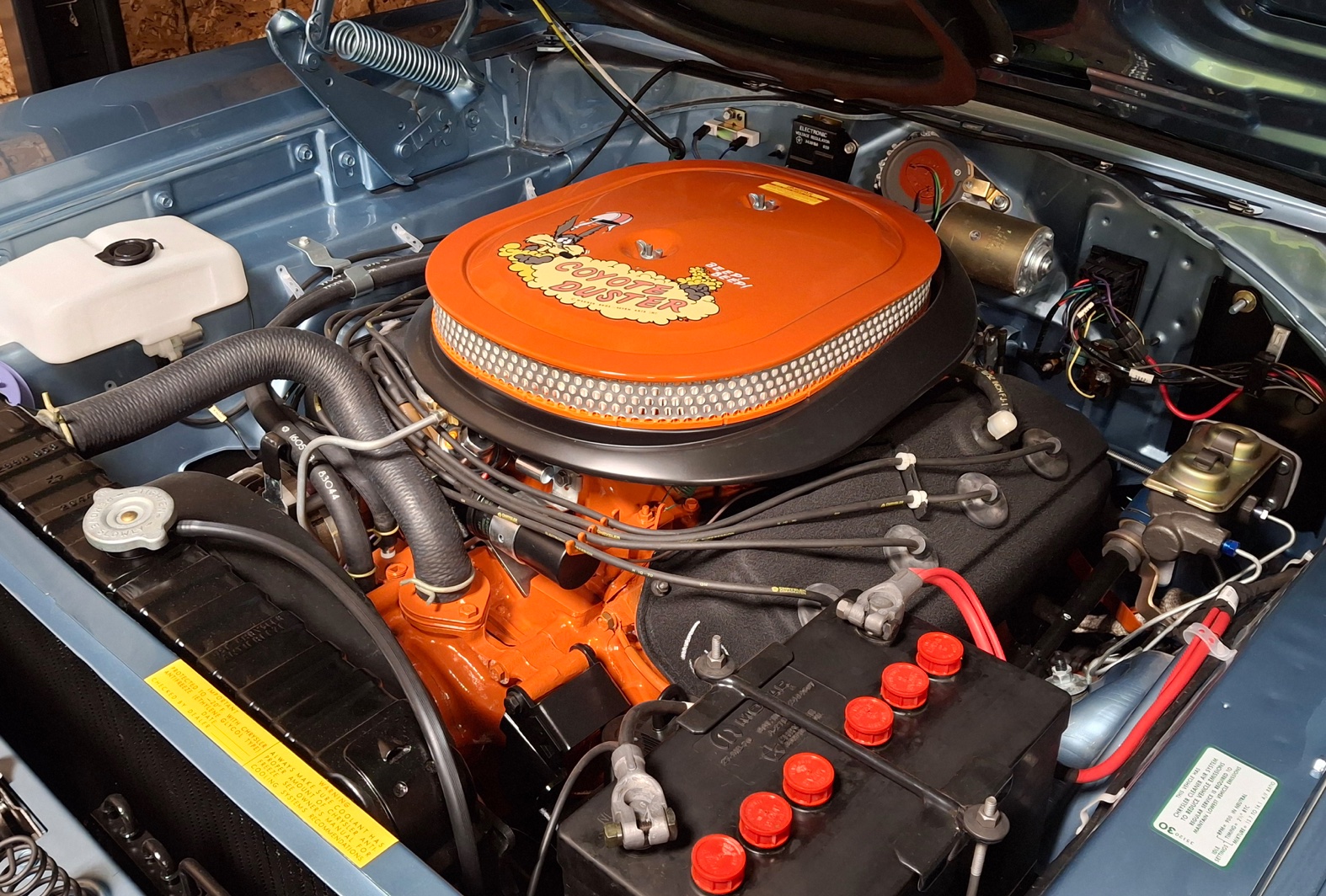A close-up view of a brightly colored, detailed car engine with an orange air filter cover labeled Six Pack, black hoses, red battery connectors, and various engine components inside a clean engine bay.