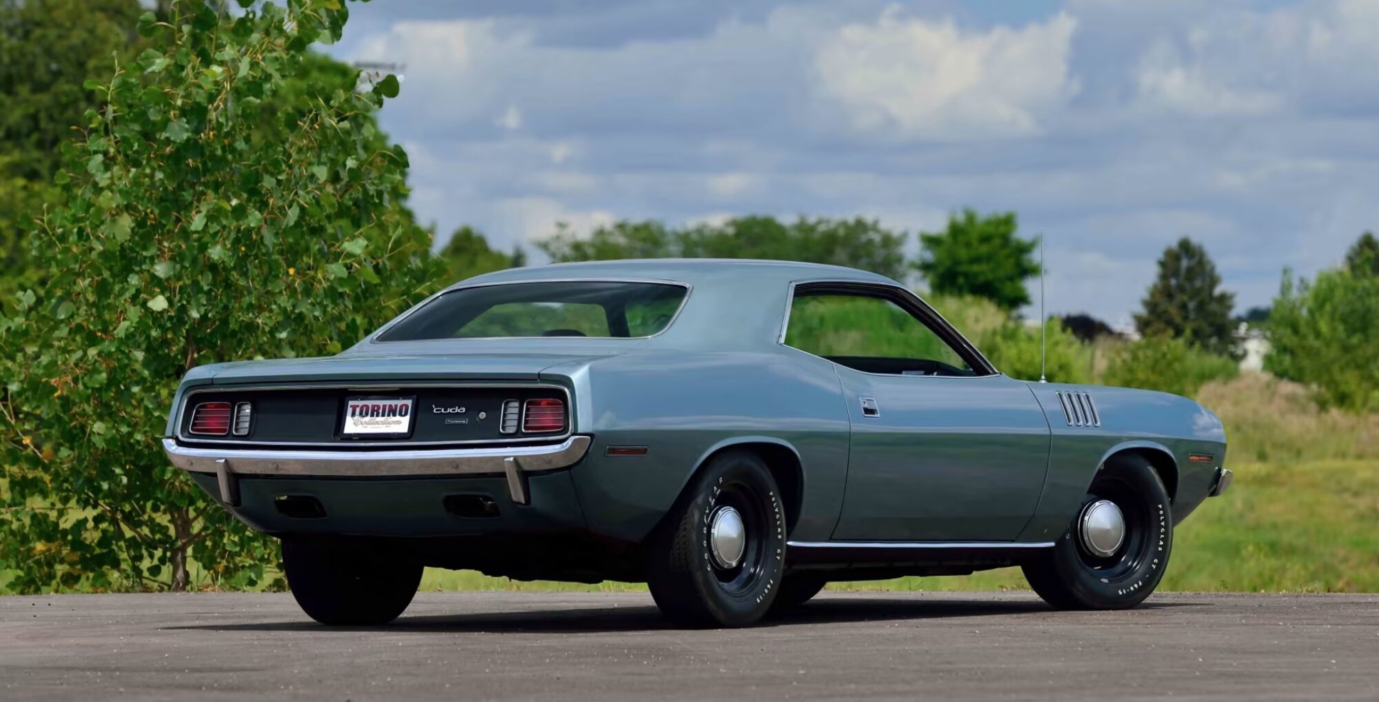 A classic silver-blue muscle car parked on pavement, viewed from the rear left side. The car has a sleek, vintage design with round taillights and black wheels. Lush green trees and a cloudy sky are in the background.