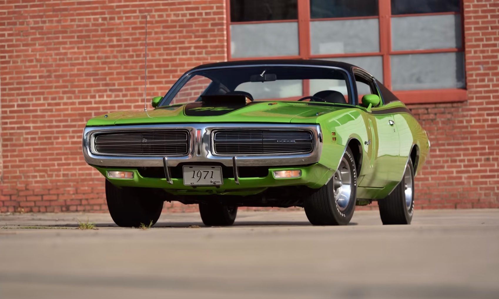 A bright green 1971 Dodge Charger classic muscle car is parked on a concrete surface in front of a red brick building with large windows.