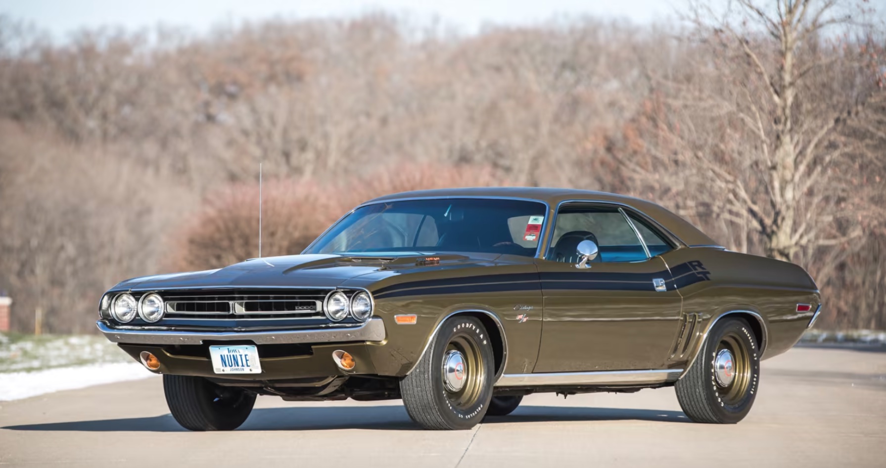 A vintage dark green 1970 Dodge Challenger muscle car is parked on a paved road with bare trees and a light dusting of snow in the background. The car has a KNATE license plate.