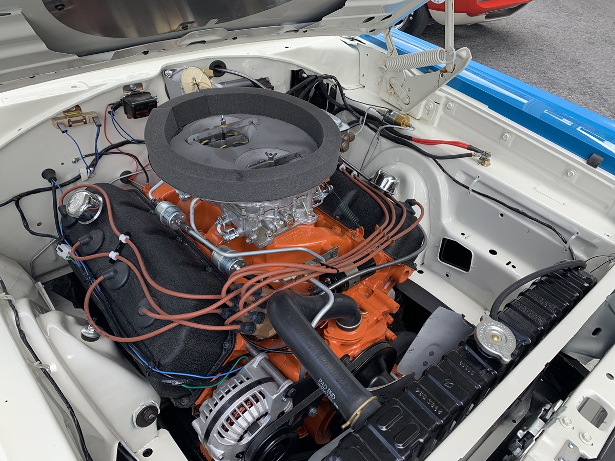Close-up view of a car’s engine bay featuring a clean, restored orange engine with black valve covers, visible wiring, hoses, and components, set inside a white-painted compartment.
