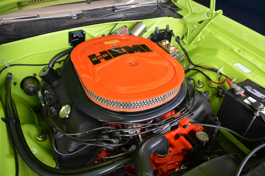 Close-up of a car engine bay featuring a bright orange air filter cover labeled 426 HEMI inside a lime green vehicle. Various engine components and battery are visible around the engine.