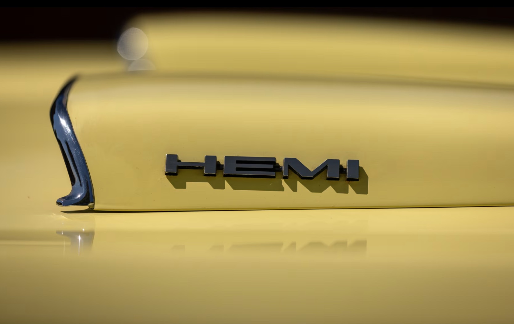 Close-up of a yellow car with a raised section on the hood displaying the black HEMI badge and a chrome accent on the left side. The background is out of focus.