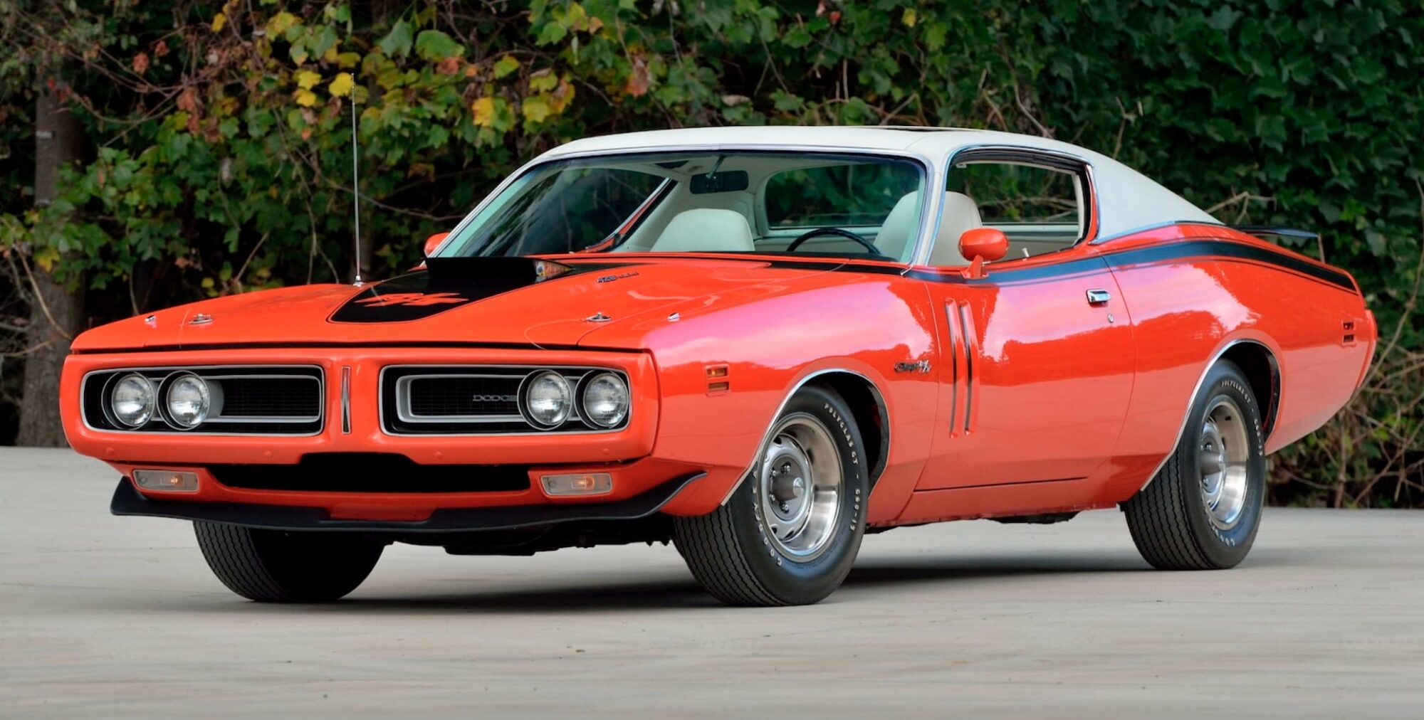 A bright orange classic Dodge Charger muscle car with a white vinyl roof and black hood scoop is parked outdoors on a concrete surface, surrounded by greenery in the background.