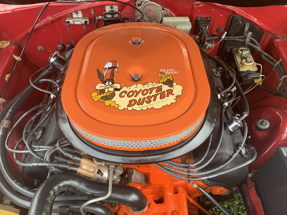 An orange air cleaner with Coyote Duster and a cartoon Road Runner is mounted on a car engine, surrounded by red bodywork and various engine components.